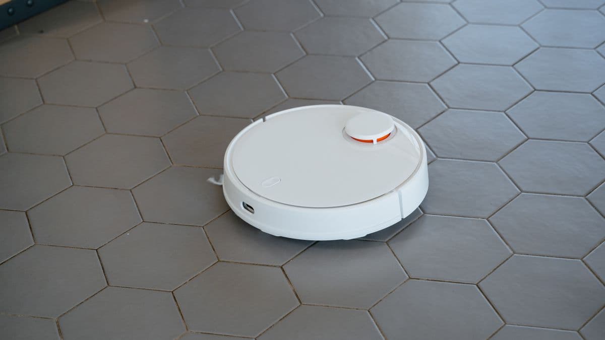 A white robot vacuum with a lidar turret cleaning grey hexagonal tiles, the only tier of household robot shipping in the tens of millions
