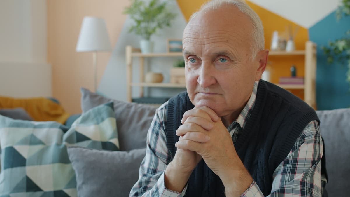 A white-haired man sits alone on a grey sofa in a bright living room with his hands clasped, the elder-care setting where the demand for home robots is most acute