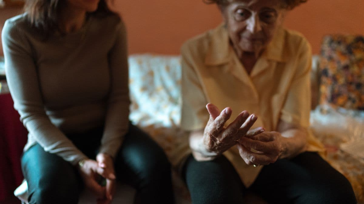 An elderly woman examining her hands while a younger caregiver sits beside her at home, the kind of care work Gates argues societies may deliberately reserve for humans