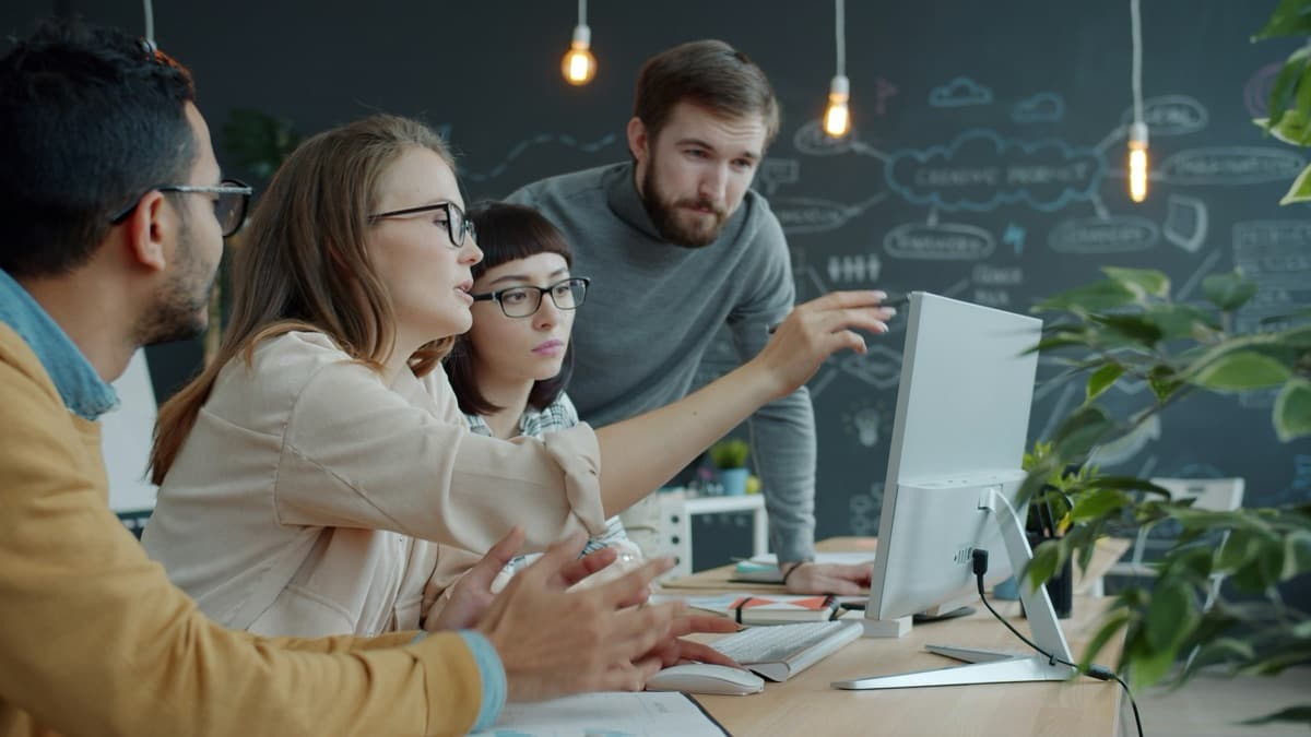 Four colleagues gathered around a monitor in a working product discussion, with hand-drawn flow diagrams on a chalkboard wall behind them