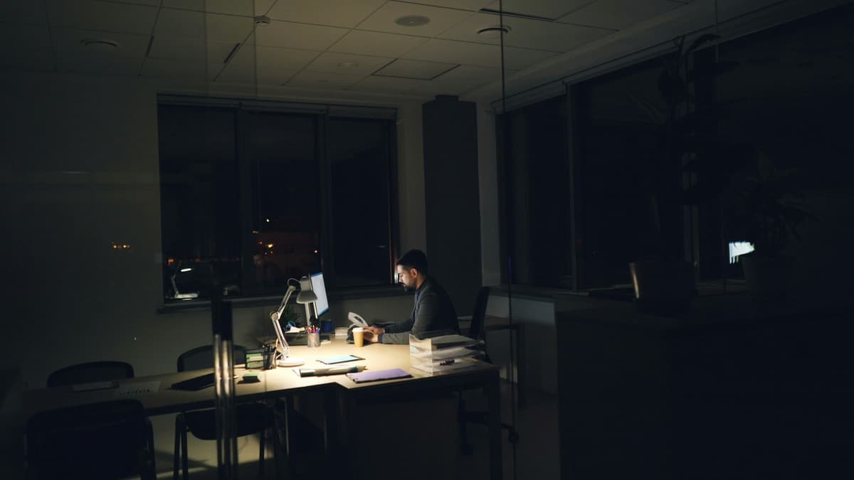 A single person working alone at a desk lamp in a dark, empty open-plan office at night, surrounded by rows of empty chairs